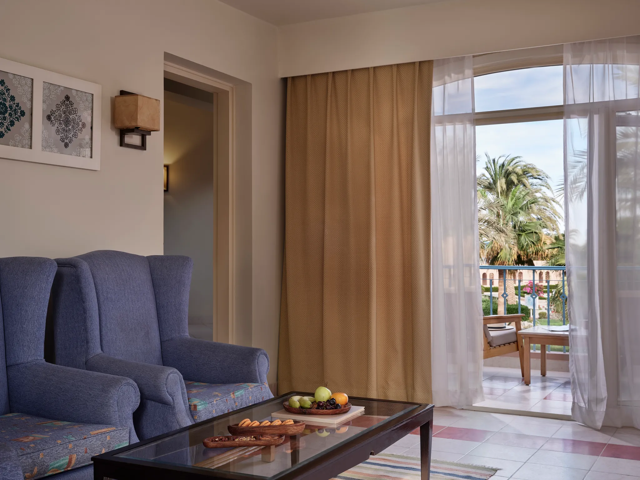 غرفة المعيشة في جناح «بارادايس» (Paradise Suite) التي تتميز بكراسي بذراعين زرقاء، وطاولة قهوة زجاجية عليها طبق فواكه، ومدخل شرفة يطل على حدائق المنتجع في فندق «كلوب باراديسيو» (Club Paradisio) بالجونة