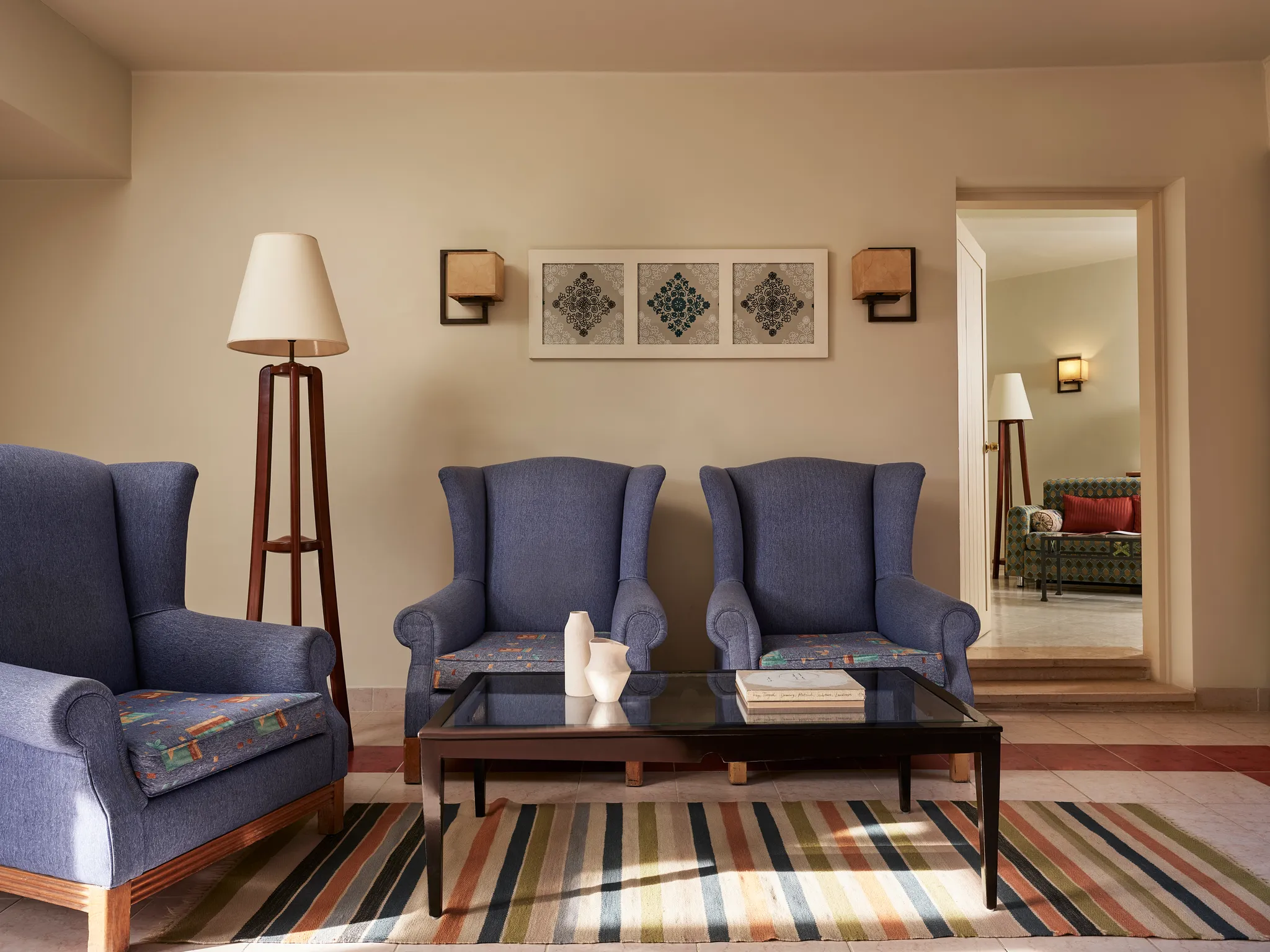 تجهيزات غرفة المعيشة الكاملة في جناح «بارادايس» (Paradise Suite) مع ثلاثة كراسي بذراعين زرقاء، ومصباح أرضي ثلاثي القوائم، وطاولة قهوة زجاجية فوق سجادة مخططة، وباب يربط الغرفة بالغرفة المجاورة في فندق «كلوب باراديسيو» (Club Paradisio) بالجونة.