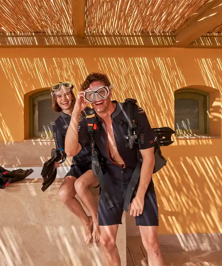 A woman and a man preparing to dive, wearing diving equipment and adjusting their gear. They are at a diving platform or shore, ready for their underwater adventure.