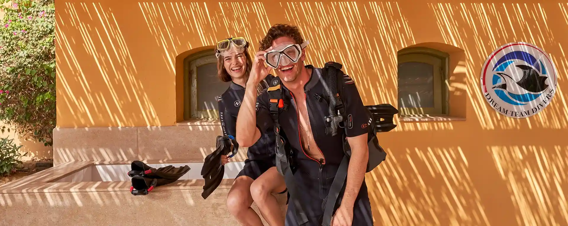 A woman and a man preparing to dive, wearing diving equipment and adjusting their gear. They are at a diving platform or shore, ready for their underwater adventure.