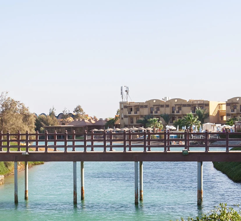 The Sultan Bey Bridge spanning over calm water, with its elegant arches and wooden decking visible. The bridge offers a scenic view of the surrounding area, with reflections of the bridge and nearby greenery mirrored in the water below. The overall setting is tranquil and picturesque.