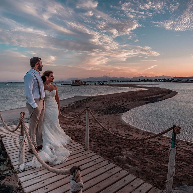 Groom and bride enjoying a beautiful sunset, standing together and embracing as the sun sets on the horizon. The scene captures a romantic and intimate moment with warm, golden hues filling the sky and creating a picturesque backdrop for the couple.