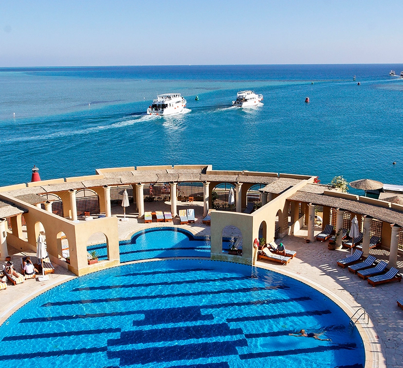 The poolside meets the seaside. A beautiful view with yachts sailing in the sea and lounges under colorful umbrellas by the pool.