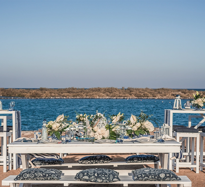 A beachfront wedding setting, with an elegantly decorated archway made of white fabric and floral arrangements, set up on the sand. Rows of white chairs are arranged facing the ocean, with a pathway leading to the altar. The scene includes a backdrop of the sea and sky, creating a romantic and picturesque atmosphere for the ceremony.