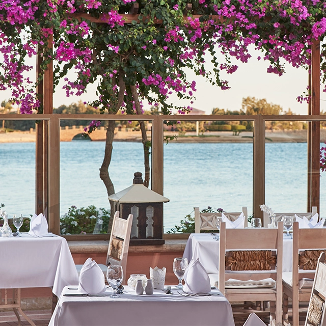 The dining area at Sultan Bey, set with neatly arranged tables and chairs under a shaded canopy. The space features a mix of indoor and outdoor seating, with a view of the surrounding lush garden and water features. Tables are elegantly set with white tablecloths, cutlery, and glassware, creating a sophisticated dining atmosphere.