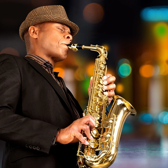 A saxophone player performing on stage, dressed in formal attire. The musician is focused on playing the saxophone, with the instrument positioned close to their mouth. The background includes dim stage lighting and possibly a blurred audience, creating an intimate and lively atmosphere.