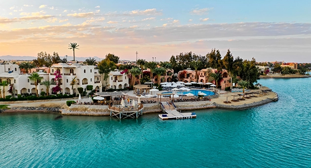 A picturesque view of Sultan Bey Beach, featuring soft golden sand and clear turquoise water. The beach is lined with sunbeds and umbrellas, with gentle waves lapping at the shore. Palm trees and lush greenery provide a tropical backdrop, enhancing the serene and relaxing atmosphere of the beach.
