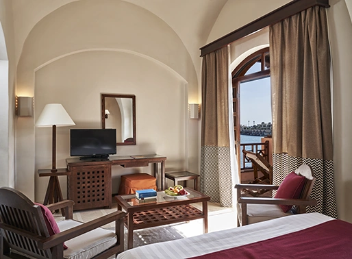 Room with a lagoon view at Red Sea, Egypt, featuring a comfortable sitting area with a television and an open balcony. The space includes stylish furnishings for relaxation and large sliding doors leading to the balcony, offering scenic views of the lagoon.