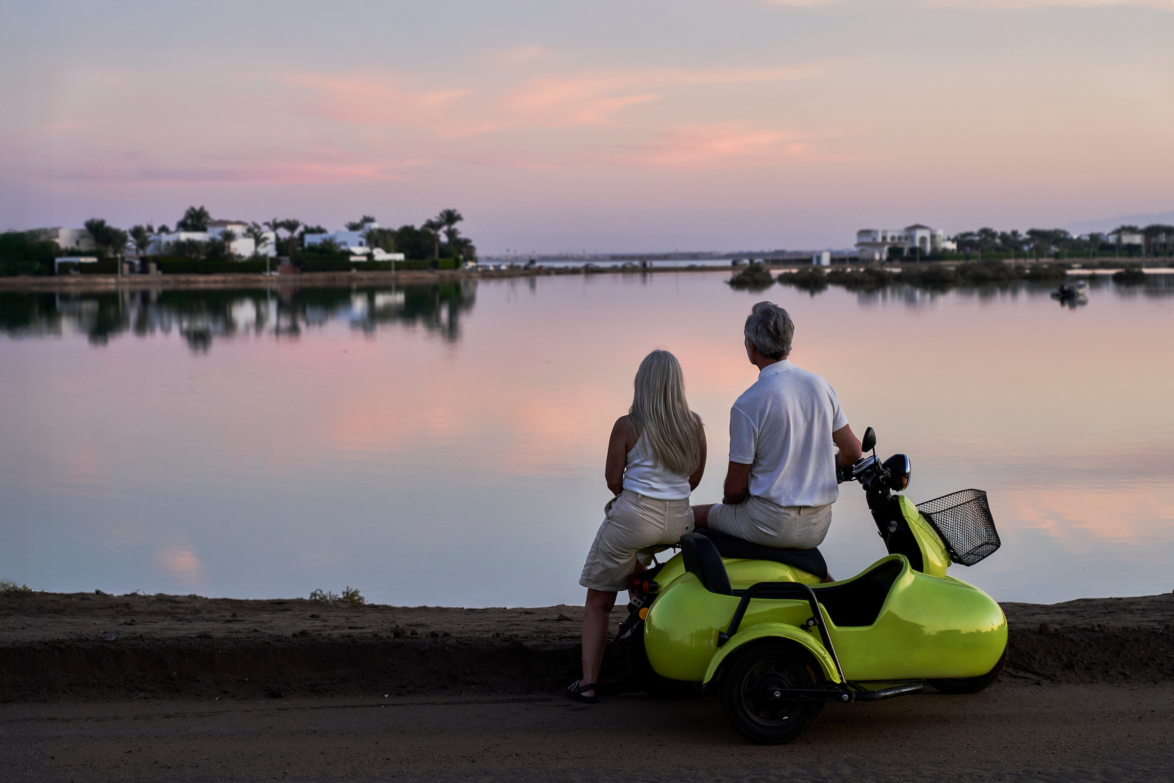 A couple sits on a bright green scooter parked beside a calm lagoon at sunset, watching the pastel sky reflect over the water with villas in the distance.
